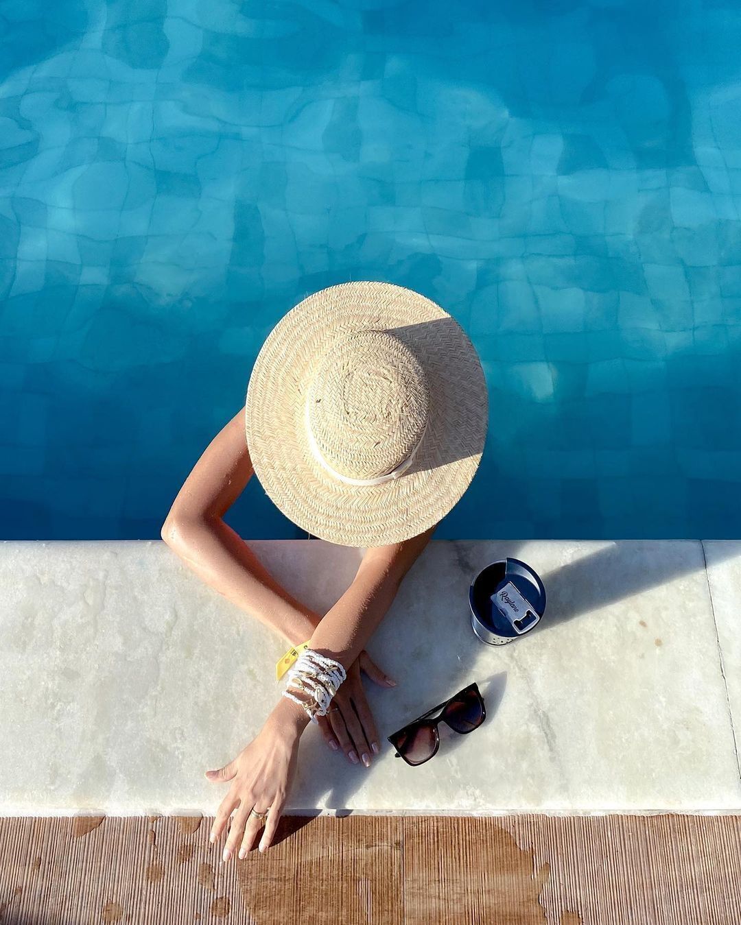Eine Person entspannt am Pool, mit Strohhut, Sonnenbrille und einer Wasserflasche.