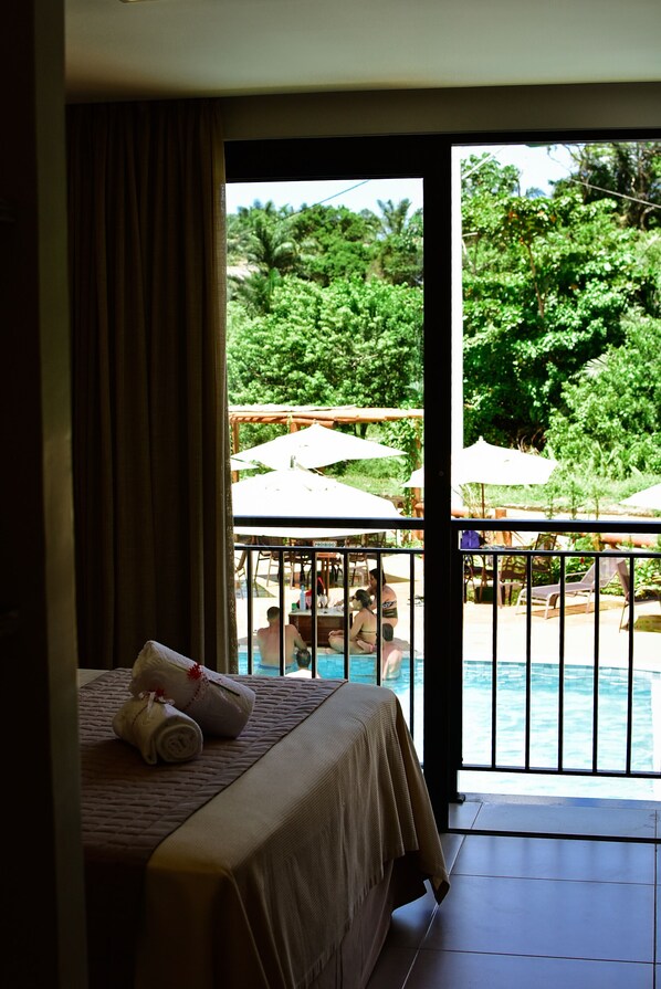 Habitación de hotel con cama y vista a un área de piscina exterior con sombrillas y sillas de descanso.