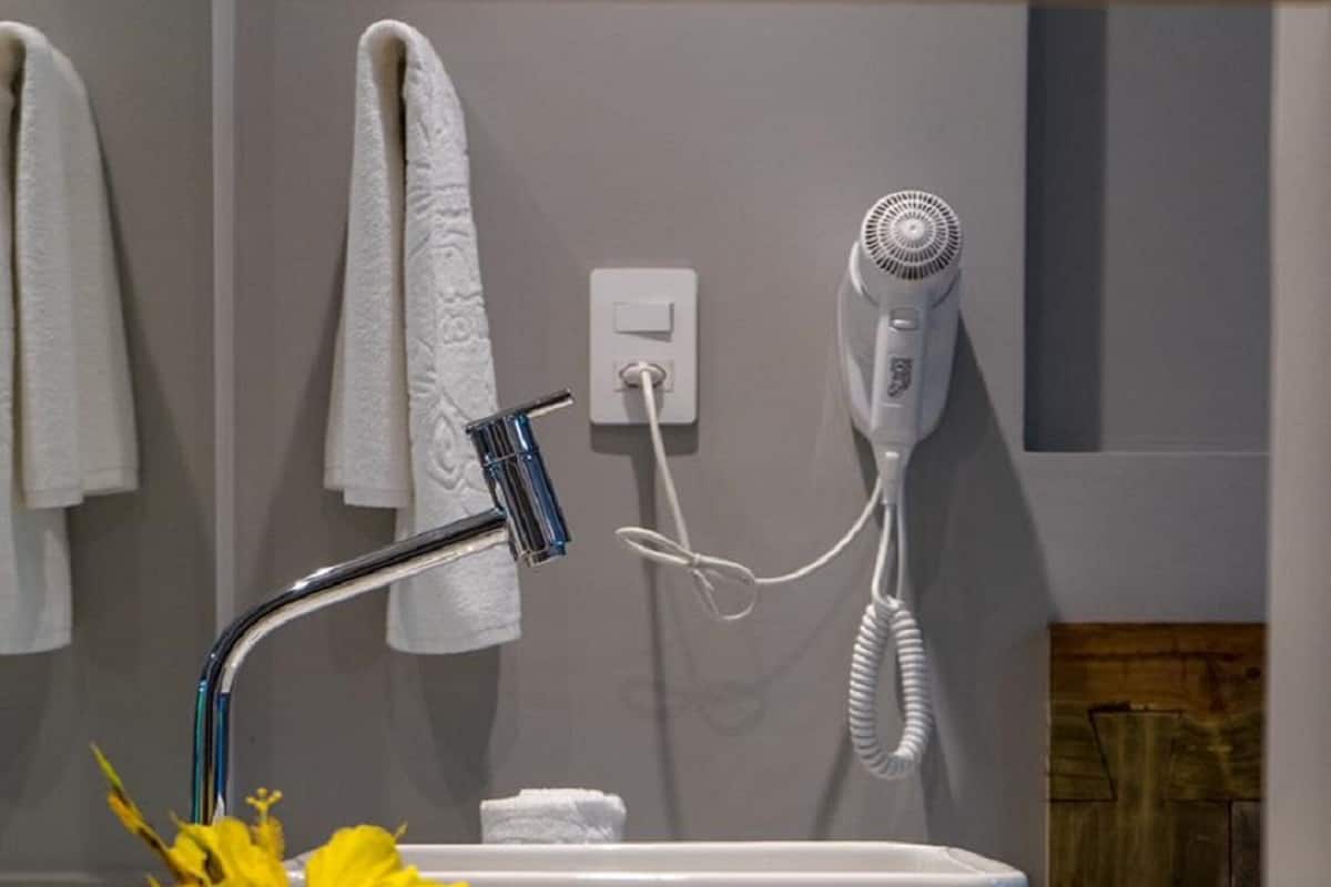 Une salle de bain avec un lavabo blanc, un porte-serviettes, un sèche-cheveux et un petit vase contenant des fleurs jaunes.