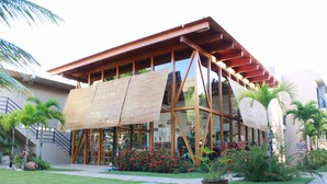 Un edificio moderno con fachada de madera y grandes ventanales, rodeado de un jardín cuidado con palmeras y flores rojas.