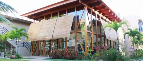 A modern building with a wooden exterior and large glass windows, surrounded by a well-maintained garden with palm trees and red flowers.