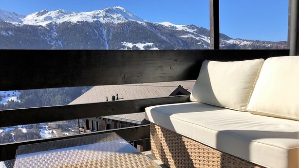 Un balcon avec un canapé en osier et une table en verre, donnant sur un paysage montagneux enneigé.