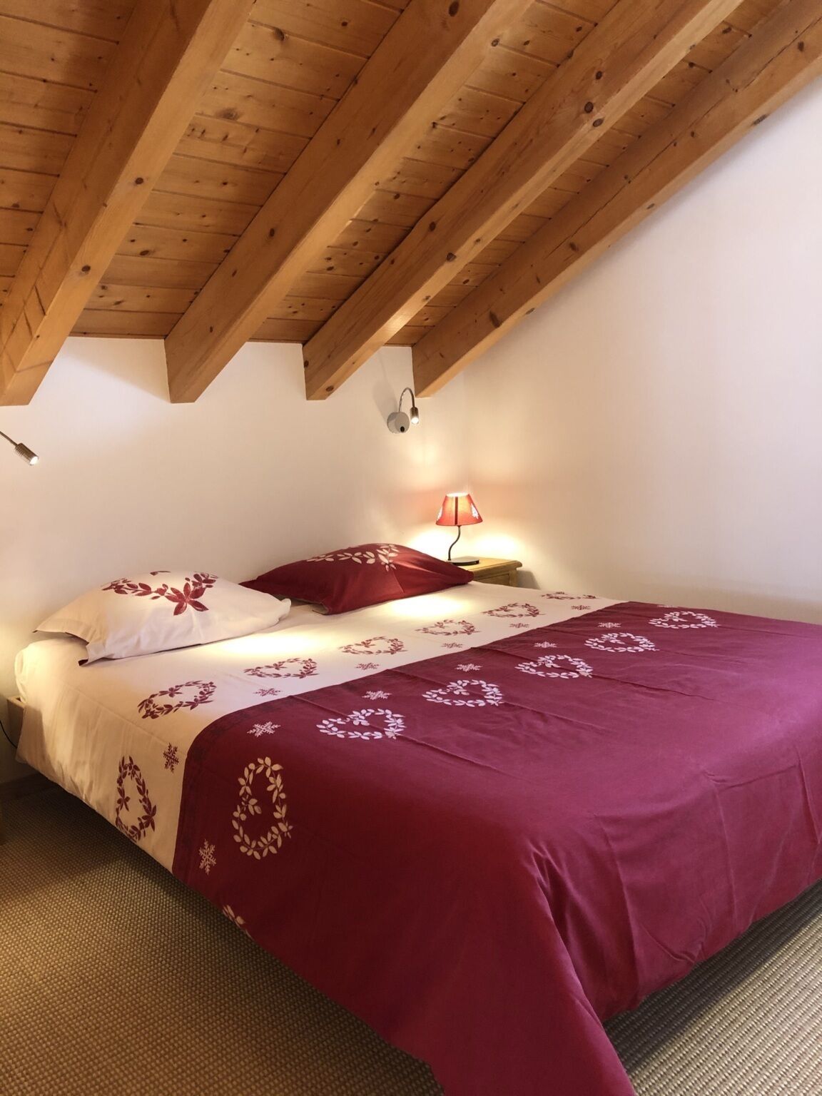 A bedroom with a wooden ceiling, a bed with a red and white bedspread, a nightstand, and a wall-mounted lamp.