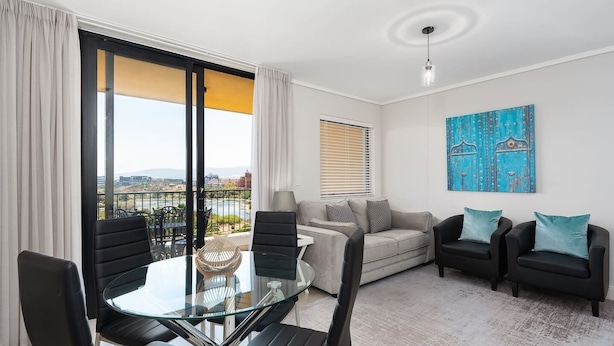 Un salón moderno con una mesa de comedor de vidrio, sillas negras, un sofá gris y dos sillones negros con cojines turquesa. Hay un ventanal con vistas al exterior, un cuadro en la pared y una lámpara de techo.