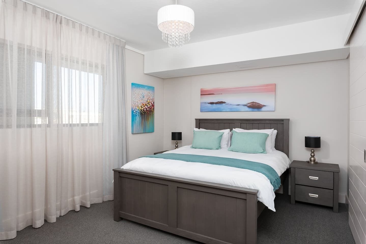 A bedroom with a bed, two bedside tables, a chandelier, and artwork on the walls.