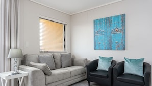 Una sala de estar con un sofá gris, dos sillones negros, una mesita de noche blanca con una lámpara, y una obra de arte azul en la pared.