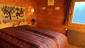 A wooden cabin room with a plaid bedspread, a mounted painting, a lamp, and a window with curtains.