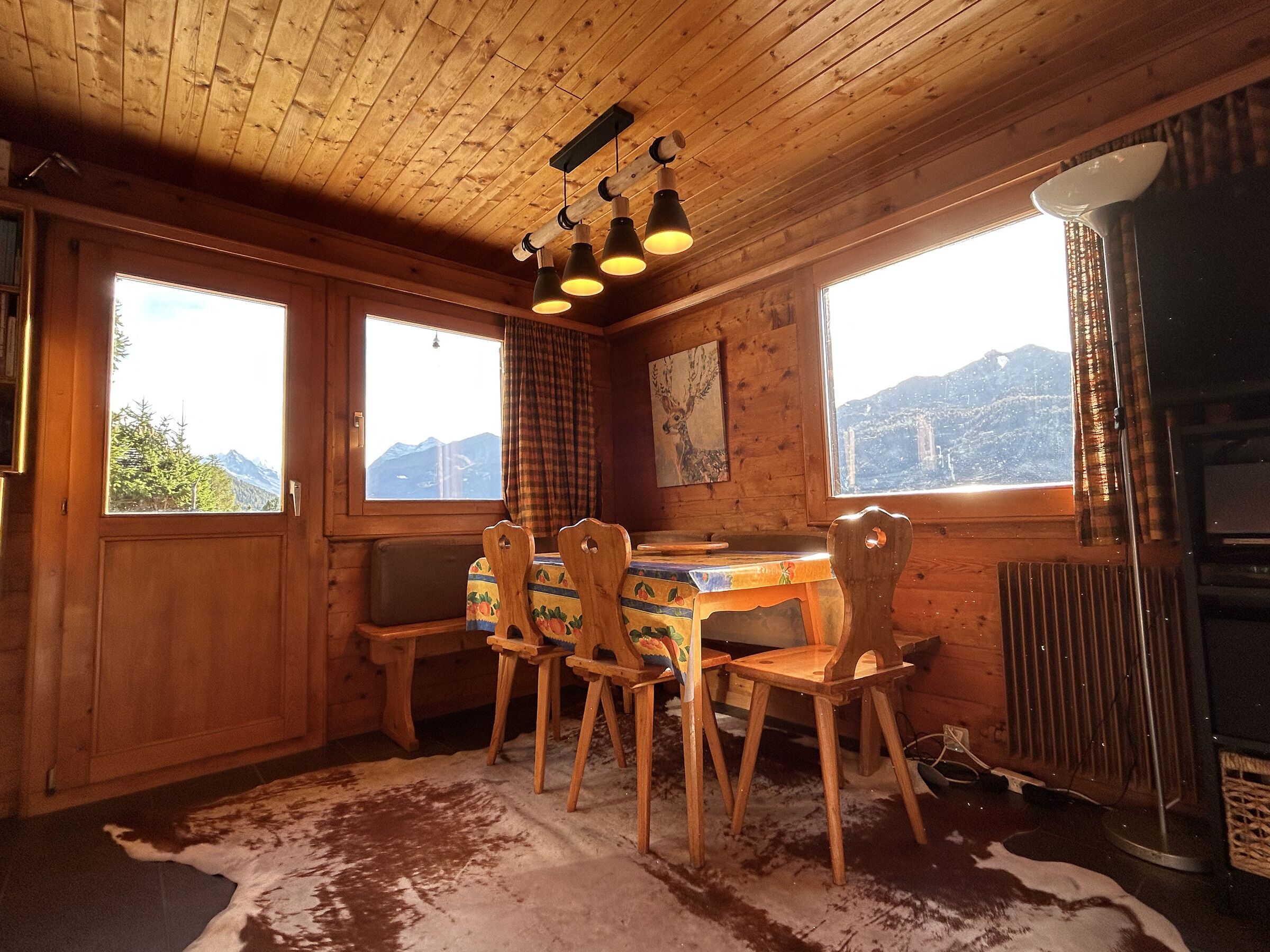 A wooden cabin interior with a dining table, chairs, and a view of mountains through large windows.