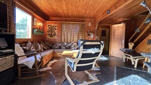 A cozy cabin interior with a wooden ceiling, a sofa, a rocking chair, a dining table, and a fireplace.