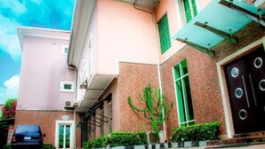 A building with a pink facade, a brick section, and a dark brown door. There is a car parked in front and a green plant in the foreground. - Villa Angelia Hotel Annex (Lagos)