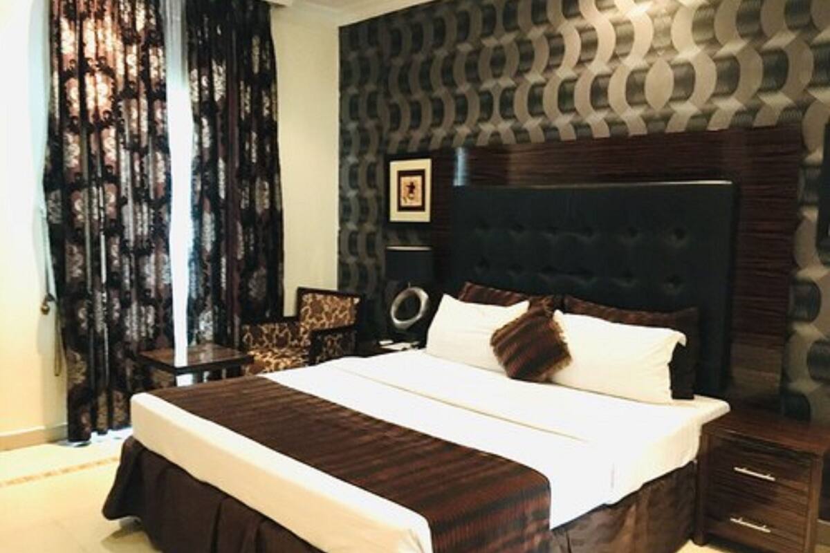A bedroom with a large bed, dark headboard, patterned wallpaper, and a curtain.