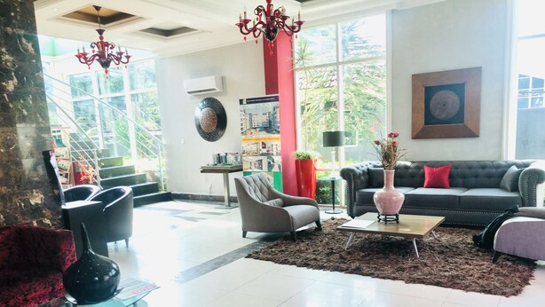 A spacious living room with a modern chandelier, a large window offering a view of greenery, a black sofa with a red pillow, and a decorative vase on a coffee table. - Villa Angelia Hotel Annex (Lagos)
