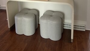 Ein Zimmer mit einem weißen Konsolentisch, zwei grauen Hocker, einem runden Spiegel und zwei an der Wand befestigten Blumenvasen.