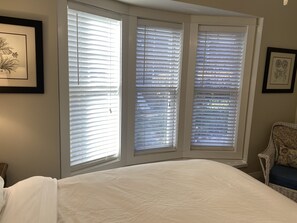 Une chambre avec un grand lit, deux fenêtres équipées de stores et une chaise.