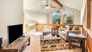 A spacious living room with a large TV, wooden furniture, a patterned rug, and a view of trees through a large window. - River Rock -- Ev # 3 Bedroom Condo by RedAwning (Estes Park)