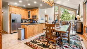A spacious kitchen with wooden cabinets, a stainless steel refrigerator, and a dining area with a wooden table and chairs. - River Rock -- Ev # 3 Bedroom Condo by RedAwning (Estes Park)