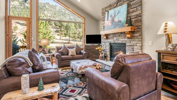 A cozy living room with leather furniture, a stone fireplace, and a large window overlooking a mountainous landscape. - River Rock -- Ev # 3 Bedroom Condo by RedAwning (Estes Park)