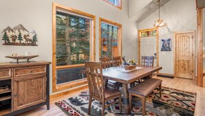 A dining area with a wooden table, chairs, and a bench. There are large windows with a view of trees and mountains. A chandelier hangs from the ceiling. - River Rock -- Ev # 3 Bedroom Condo by RedAwning (Estes Park)
