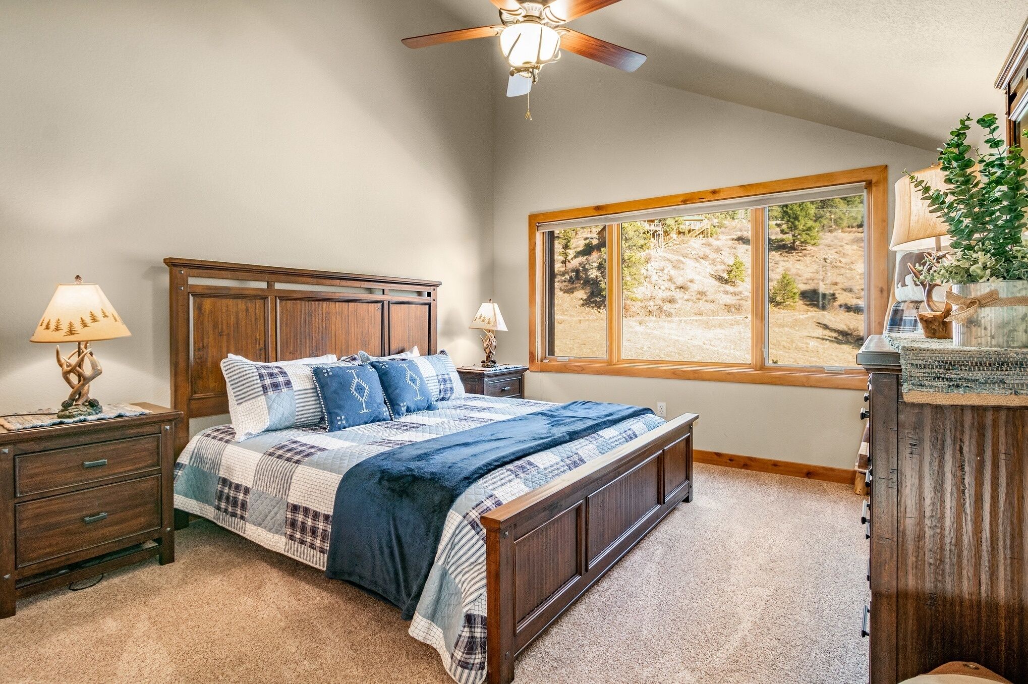 A bedroom with a large bed, wooden headboard, two bedside tables with lamps, a window with a view of a landscape, and a ceiling fan.