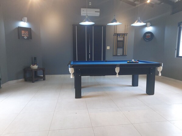 A pool table with overhead lighting, a framed picture on the wall, and a small table with a bowl of fruit.