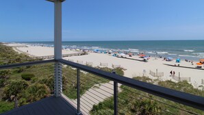 A beach view from a balcony with a railing, a wooden deck, and a clear sky.