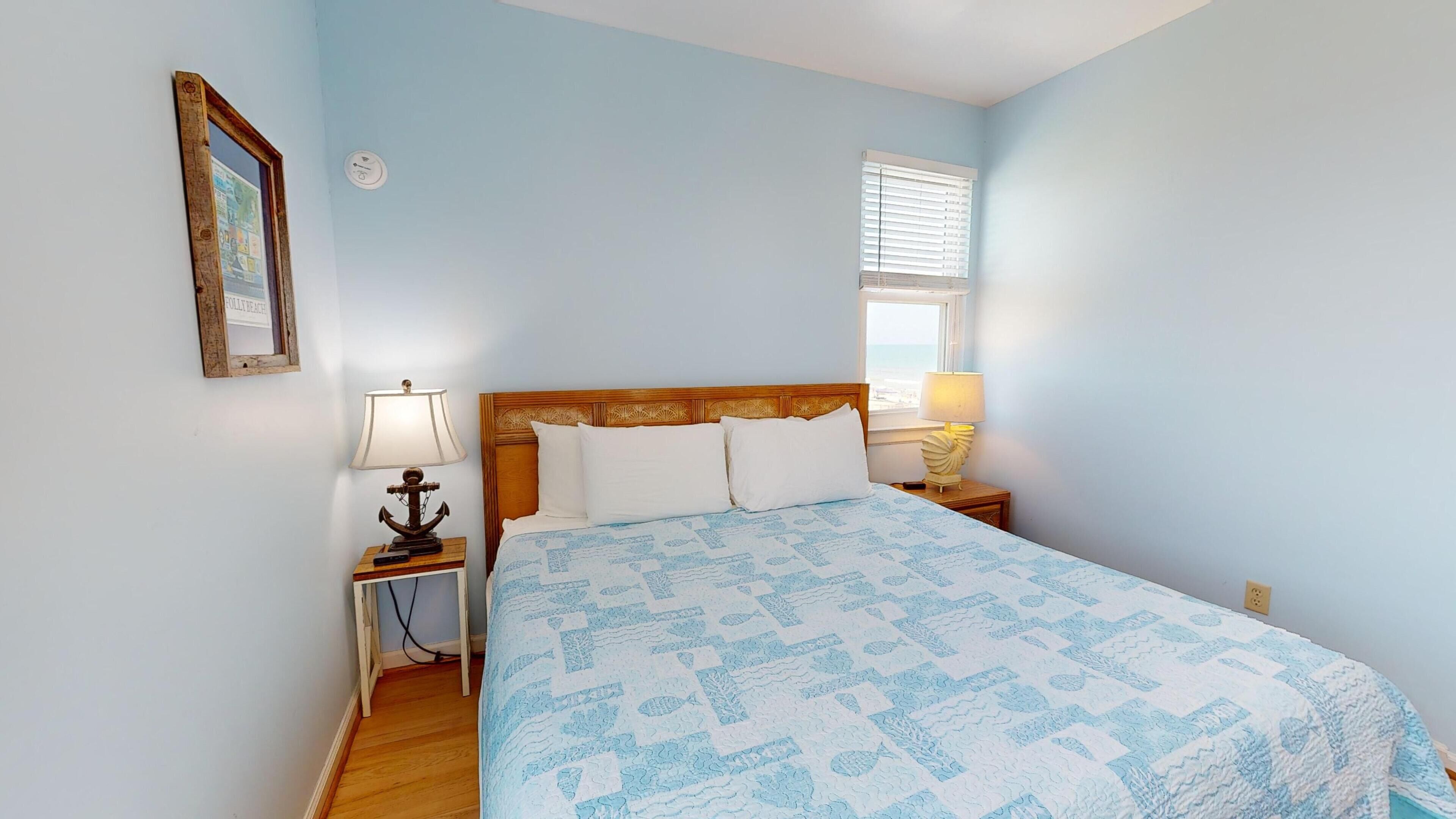 Une chambre à coucher avec une tête de lit en bois, un lit recouvert d’une couvre-lit à motifs bleus, une table de chevet avec une lampe, un interrupteur fixé au mur et une fenêtre avec des stores.