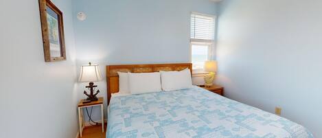 Une chambre à coucher avec une tête de lit en bois, un lit recouvert d’une couvre-lit à motifs bleus, une table de chevet avec une lampe, un interrupteur fixé au mur et une fenêtre avec des stores.