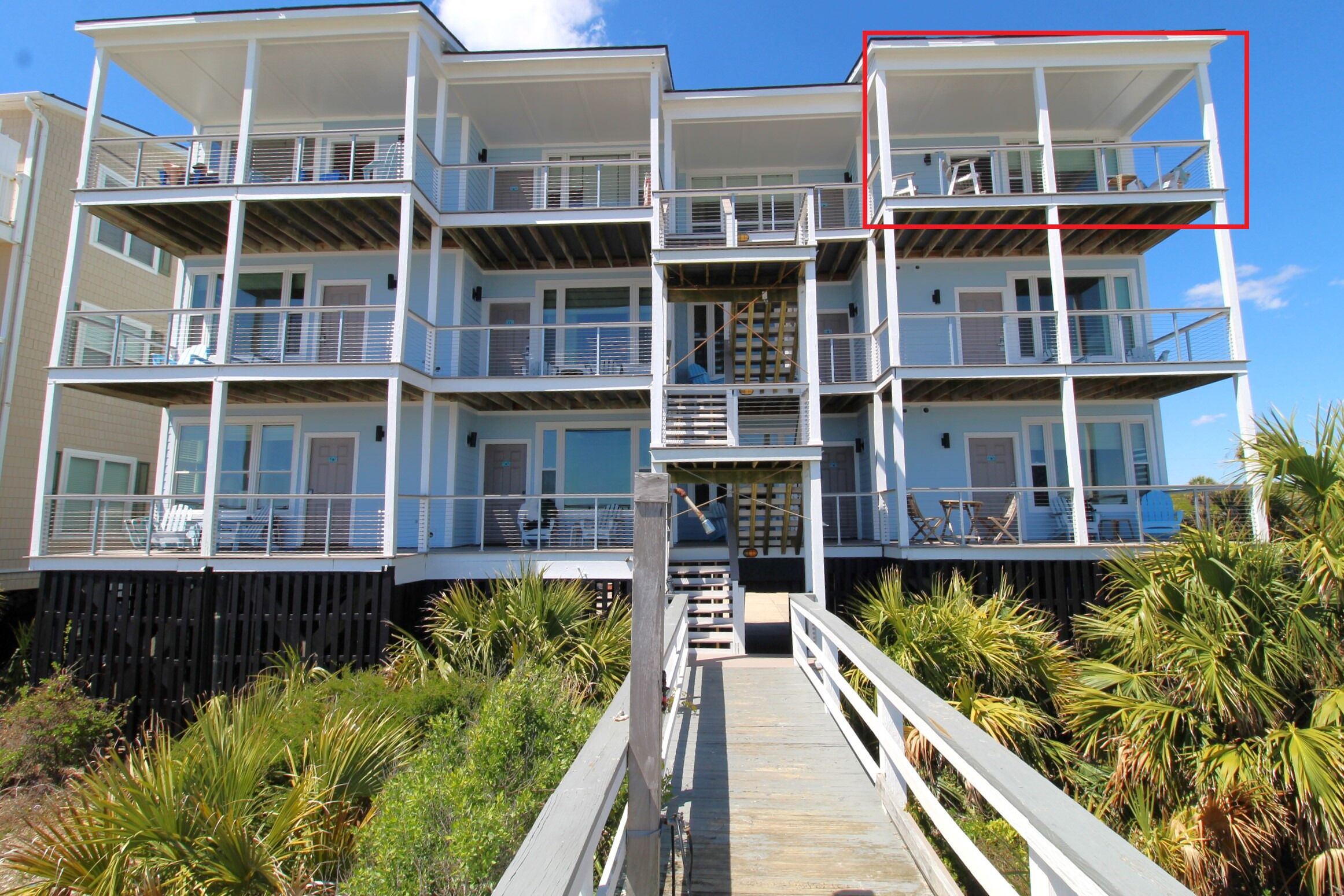 Un immeuble à plusieurs étages situé en bord de mer, doté de balcons, d’une terrasse en bois et offrant une vue sur l’océan.