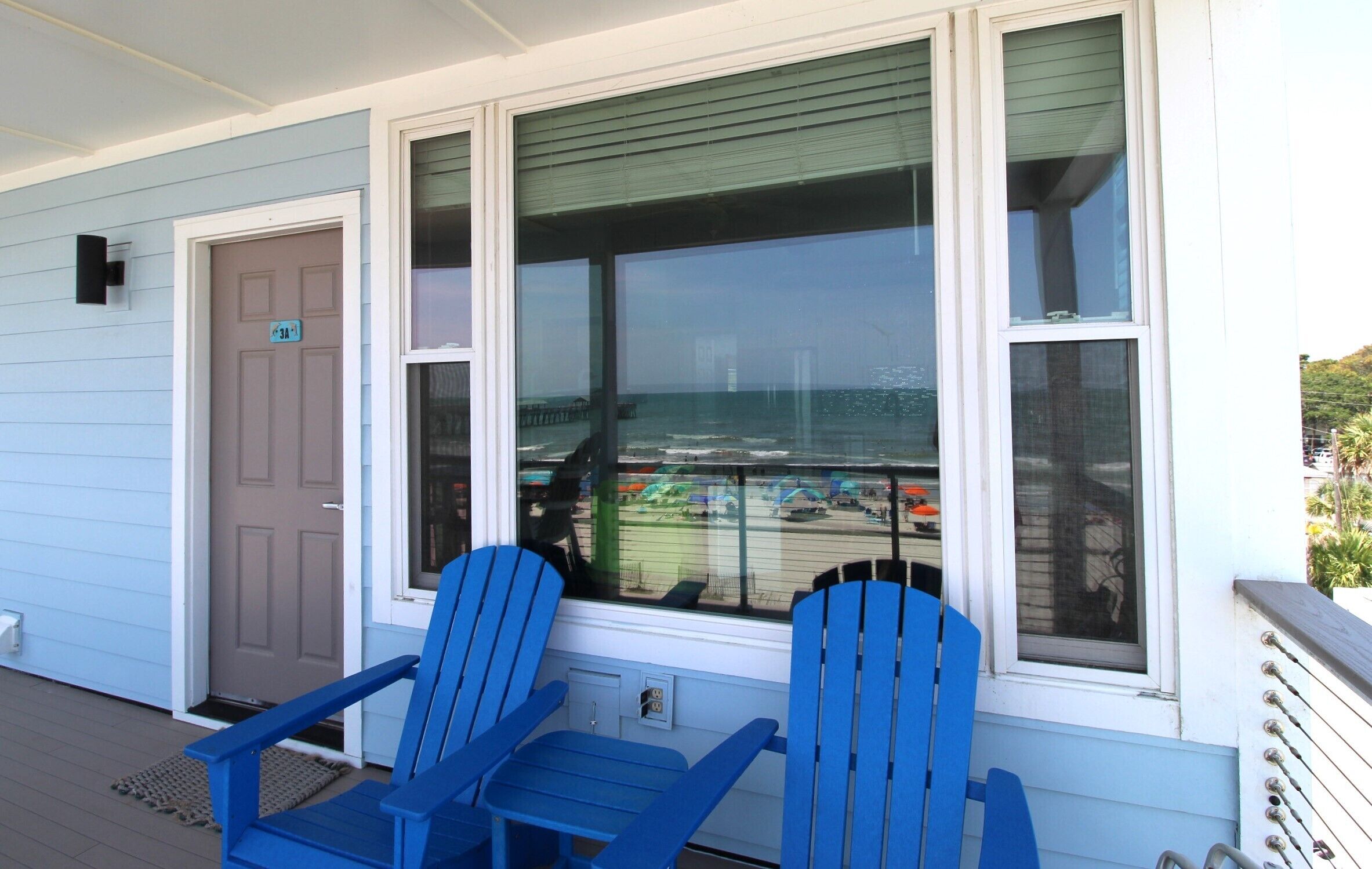 Une véranda avec deux fauteuils Adirondack bleus, une porte et une grande fenêtre donnant sur une plage où se trouvent des bateaux.
