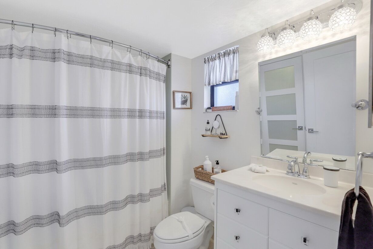 A bathroom with a shower curtain, toilet, vanity with a mirror, and a window with curtains.