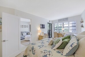 Une chambre à coucher comprenant un lit, une table de chevet, une lampe, une fenêtre avec des stores et une porte donnant sur une autre pièce.