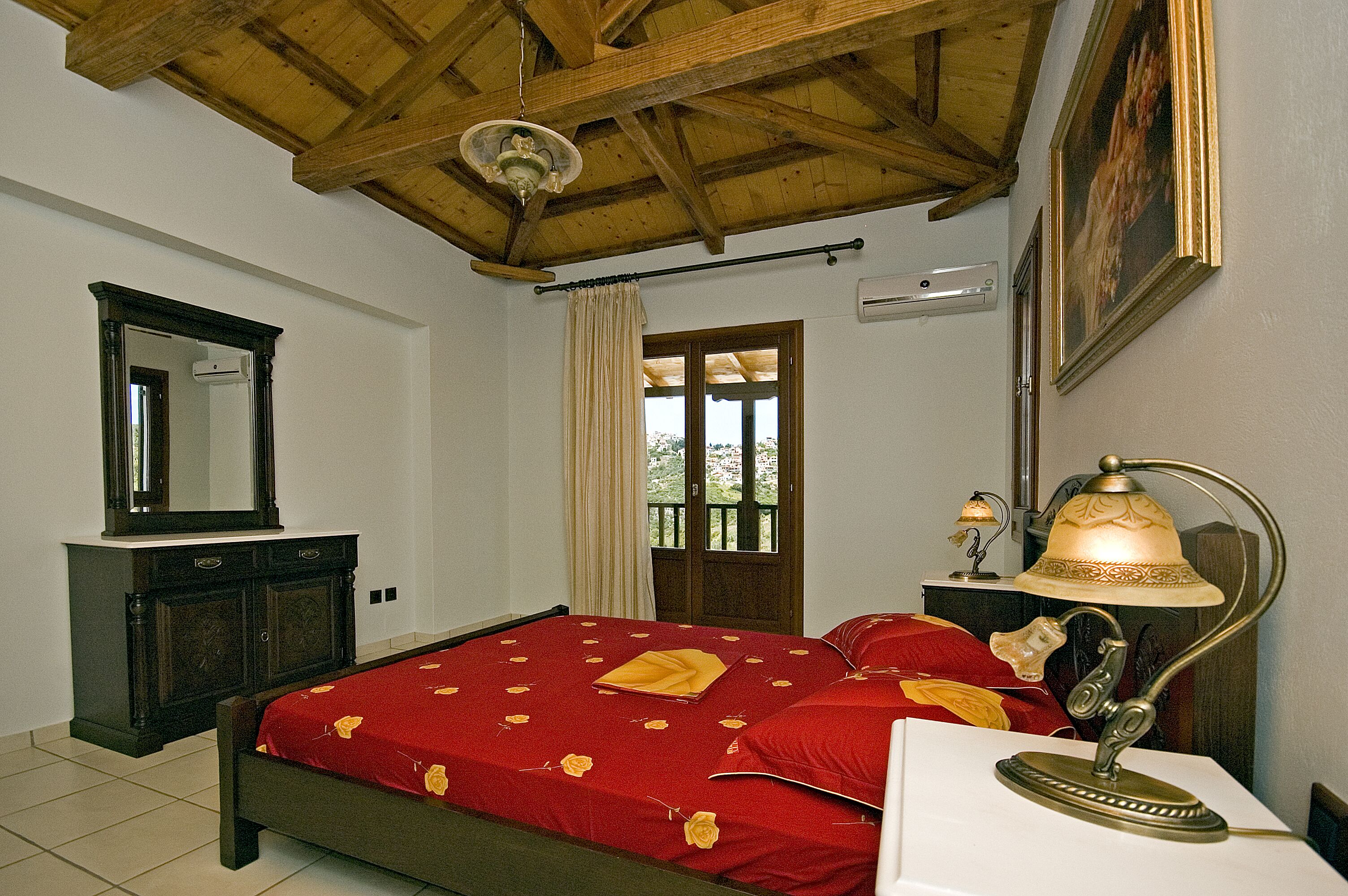 A bedroom with a wooden ceiling, a bed with red bedding, a bedside table with a lamp, a mirror, and a door leading to a balcony.