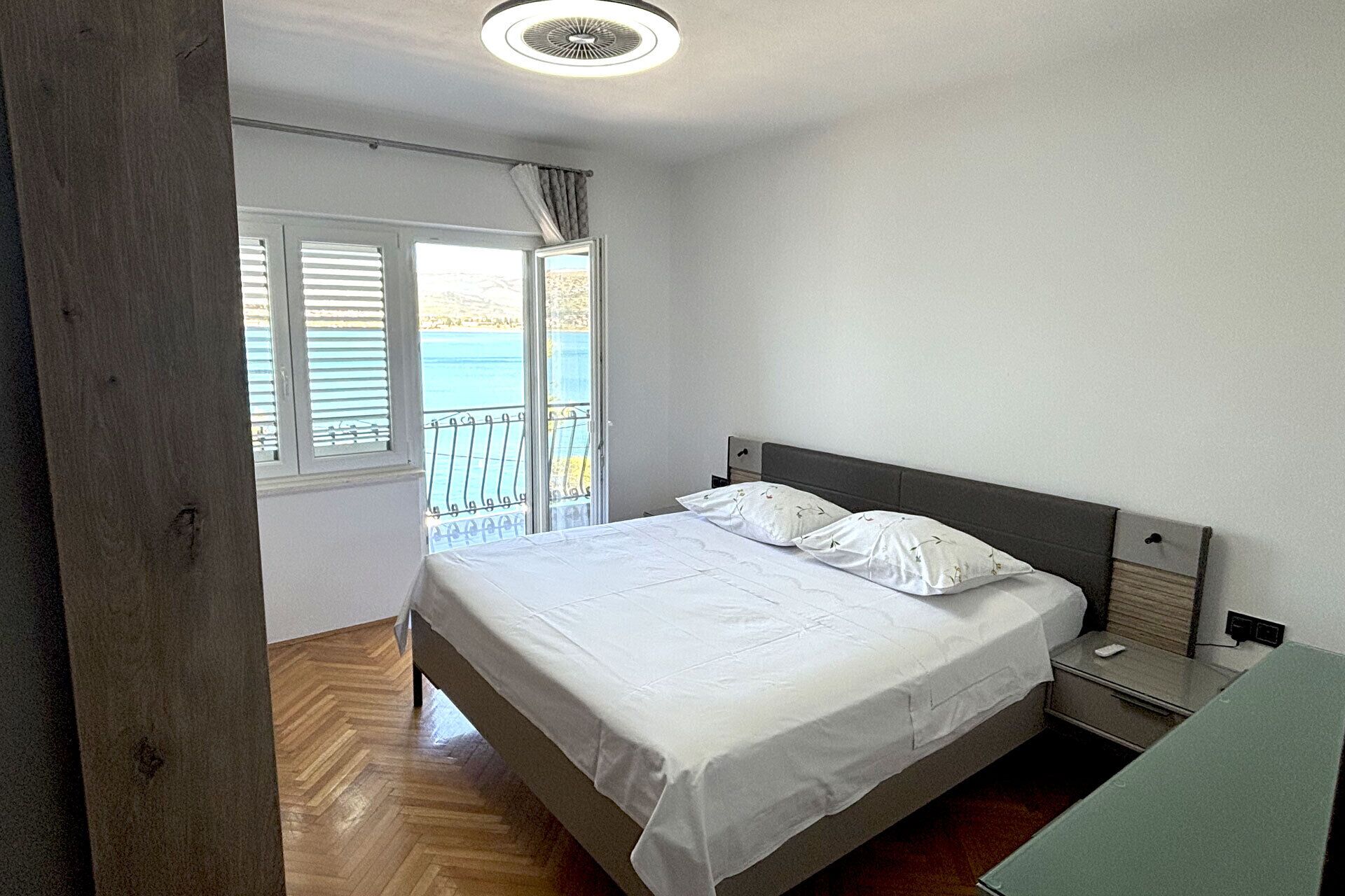 A bedroom with a bed, a nightstand, a window with blinds, and a view of the sea.