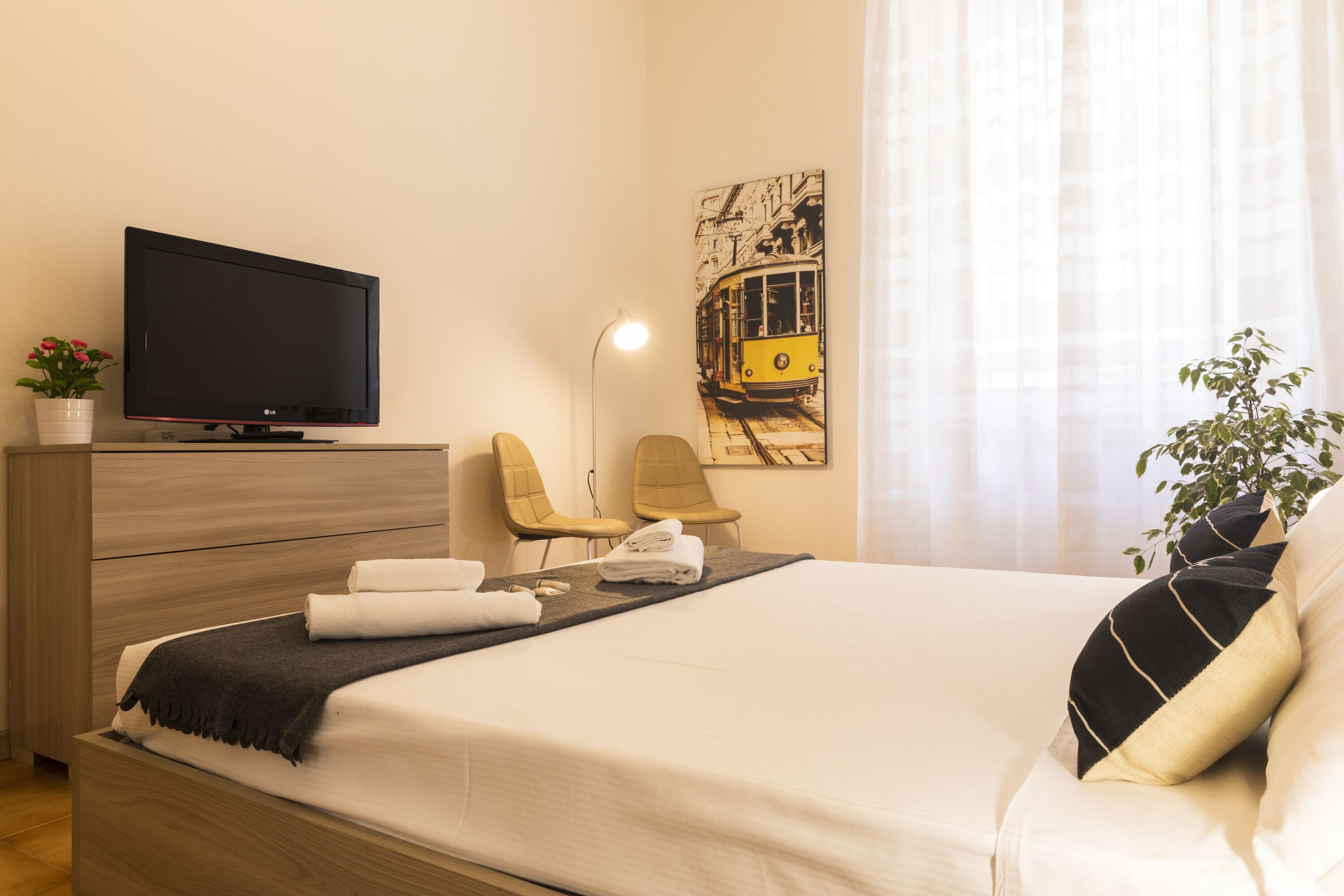 A hotel room with a bed, a TV, a desk with a lamp, two chairs, a plant, and a painting on the wall.