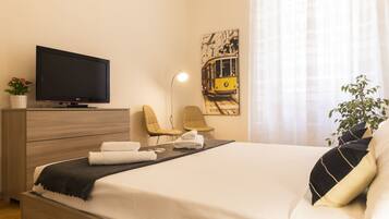 A hotel room with a bed, a TV, a desk with a lamp, two chairs, a plant, and a painting on the wall.