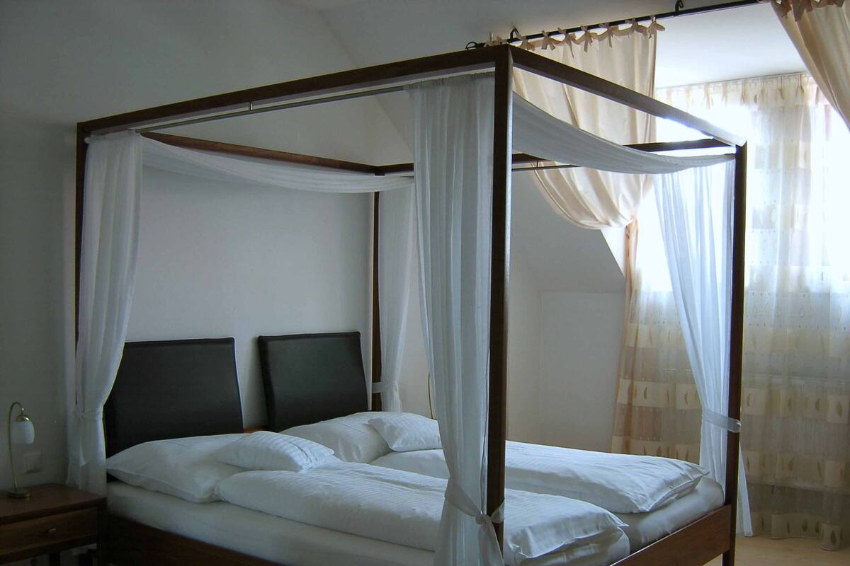 A four-poster canopy bed with white linens and sheer curtains, a bedside table with a lamp, and a window with sheer curtains.