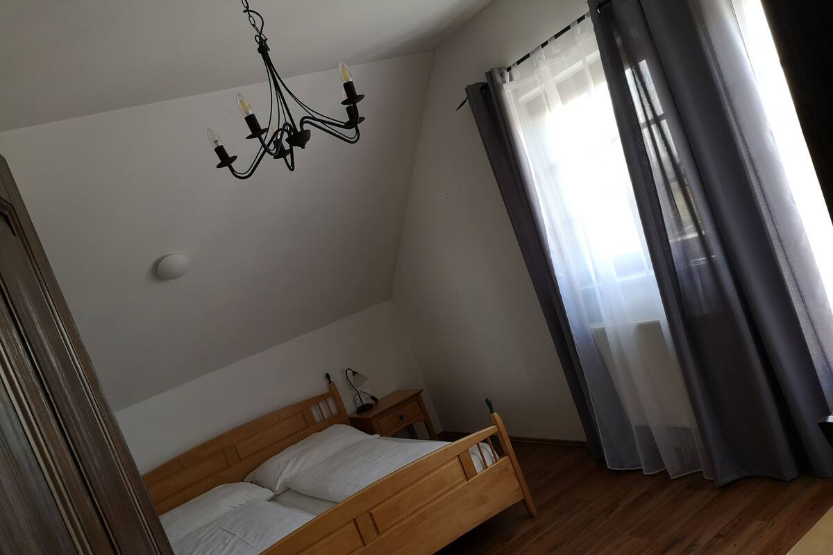 A bedroom with a wooden bed, a hanging light fixture, a bedside table, and a window with curtains.