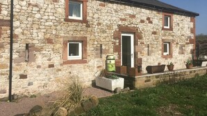 A stone building with a door, two windows, and a small garden area with potted plants. - Cosy 2-bed Cottage With Garden Near Carlisle (Carlisle)