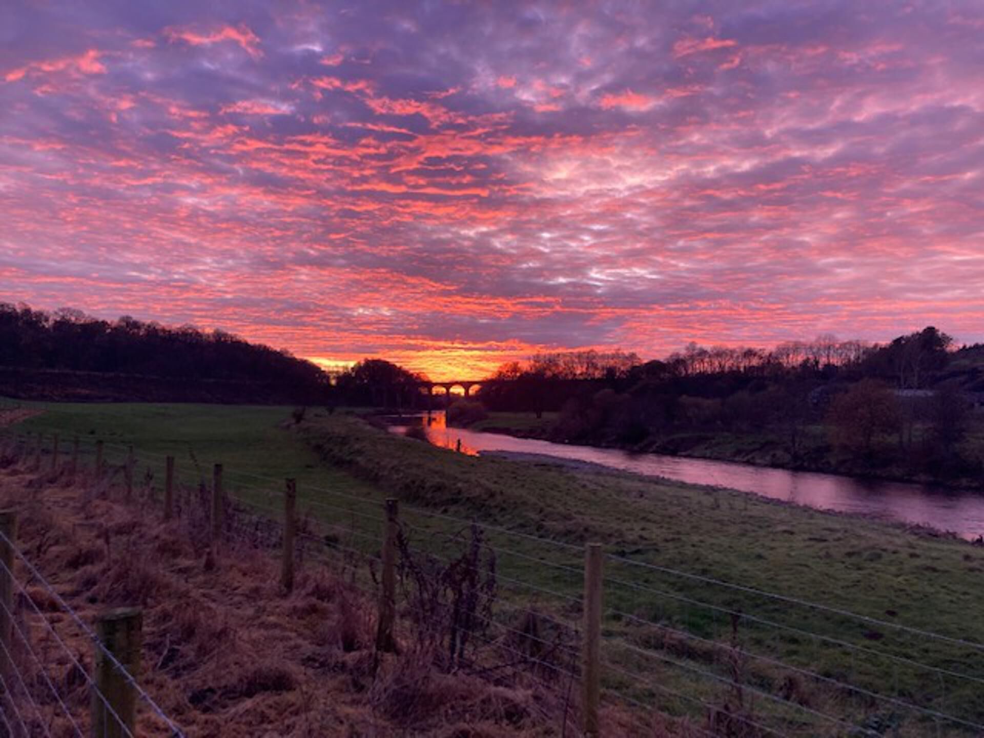 Un coucher de soleil sur une rivière, avec un pont visible au loin.
