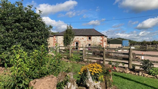 A rustic stone building with a wooden fence, surrounded by greenery and a clear sky. - Cosy 2-bed Cottage With Garden Near Carlisle (Carlisle)