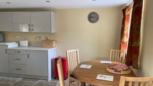 A compact kitchen with white cabinets, a red trash bin, and a wooden dining table with four chairs. - Cosy 2-bed Cottage With Garden Near Carlisle (Carlisle)