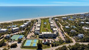 Une vue aĂ©rienne dâune station balnĂ©aire comprenant plusieurs bĂątiments, un espace piscine central, des courts de tennis et une plage en arriĂšre-plan.