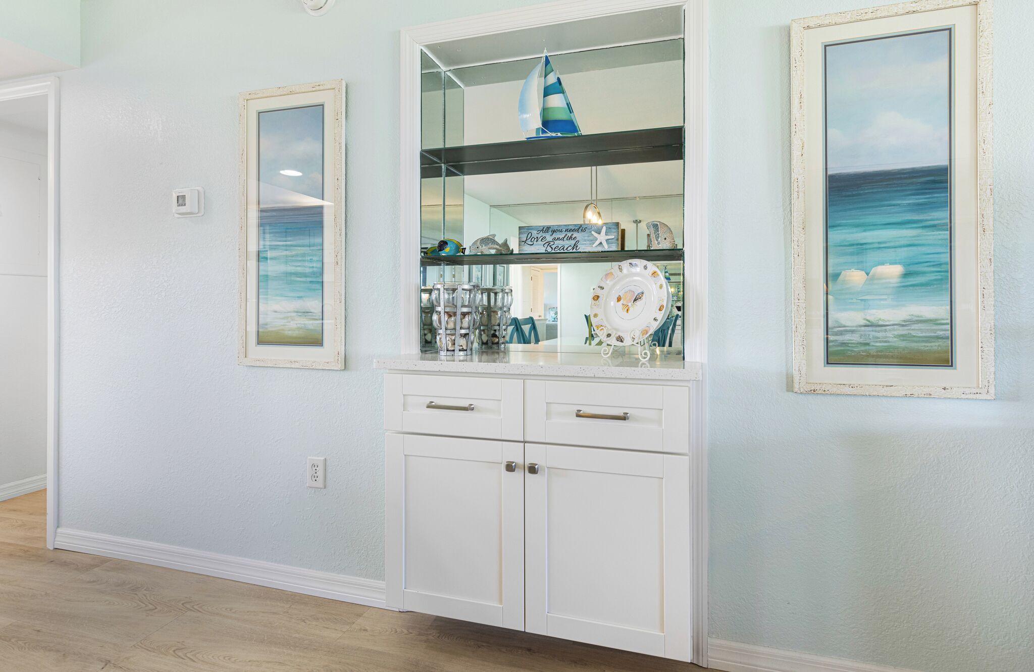 A kitchen with white cabinetry, a glass-fronted display case, and framed artwork depicting seascapes.