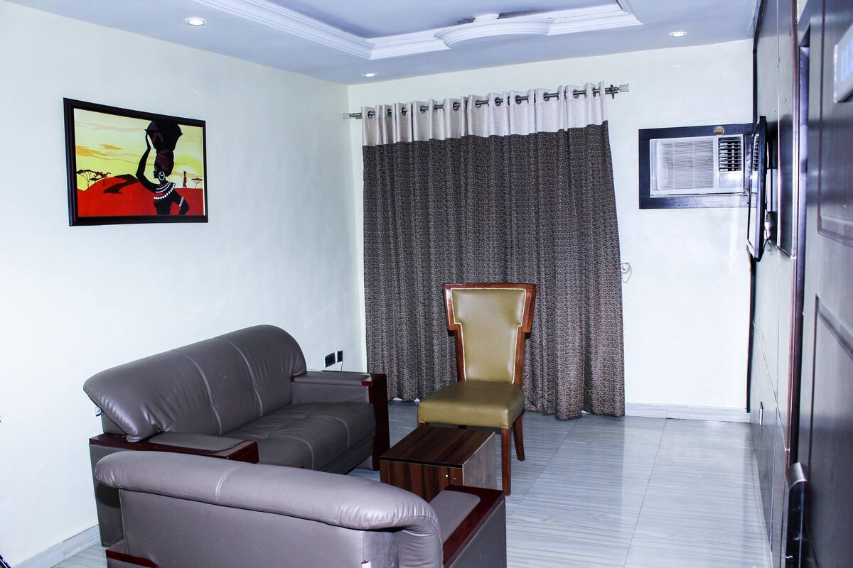 A living room with a grey sofa, a wooden coffee table, a brown armchair, a painting on the wall, and a window with curtains.
