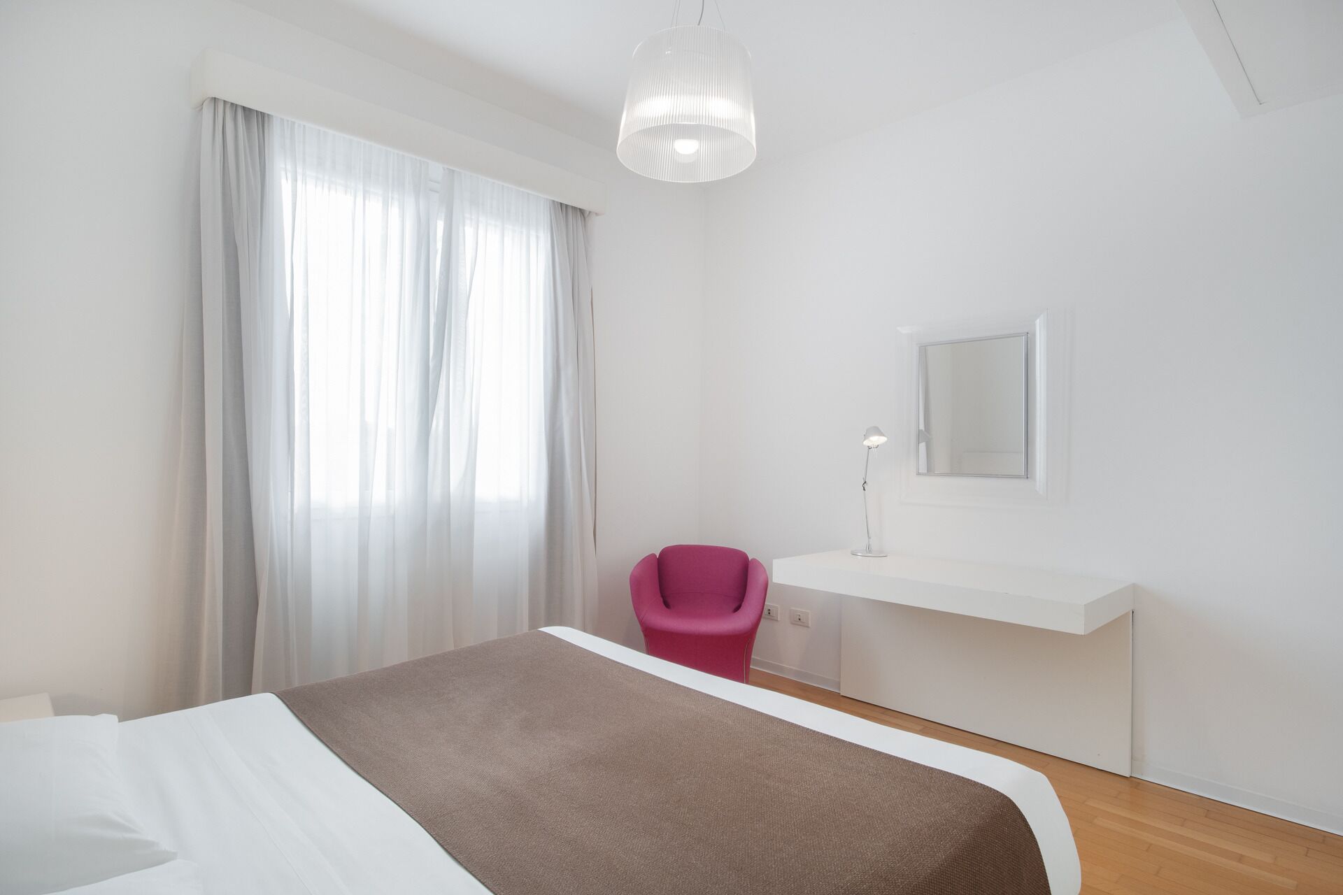 Quarto de hotel moderno com uma cama grande, uma escrivaninha com cadeira, luminária pendente e janela com cortinas translúcidas.