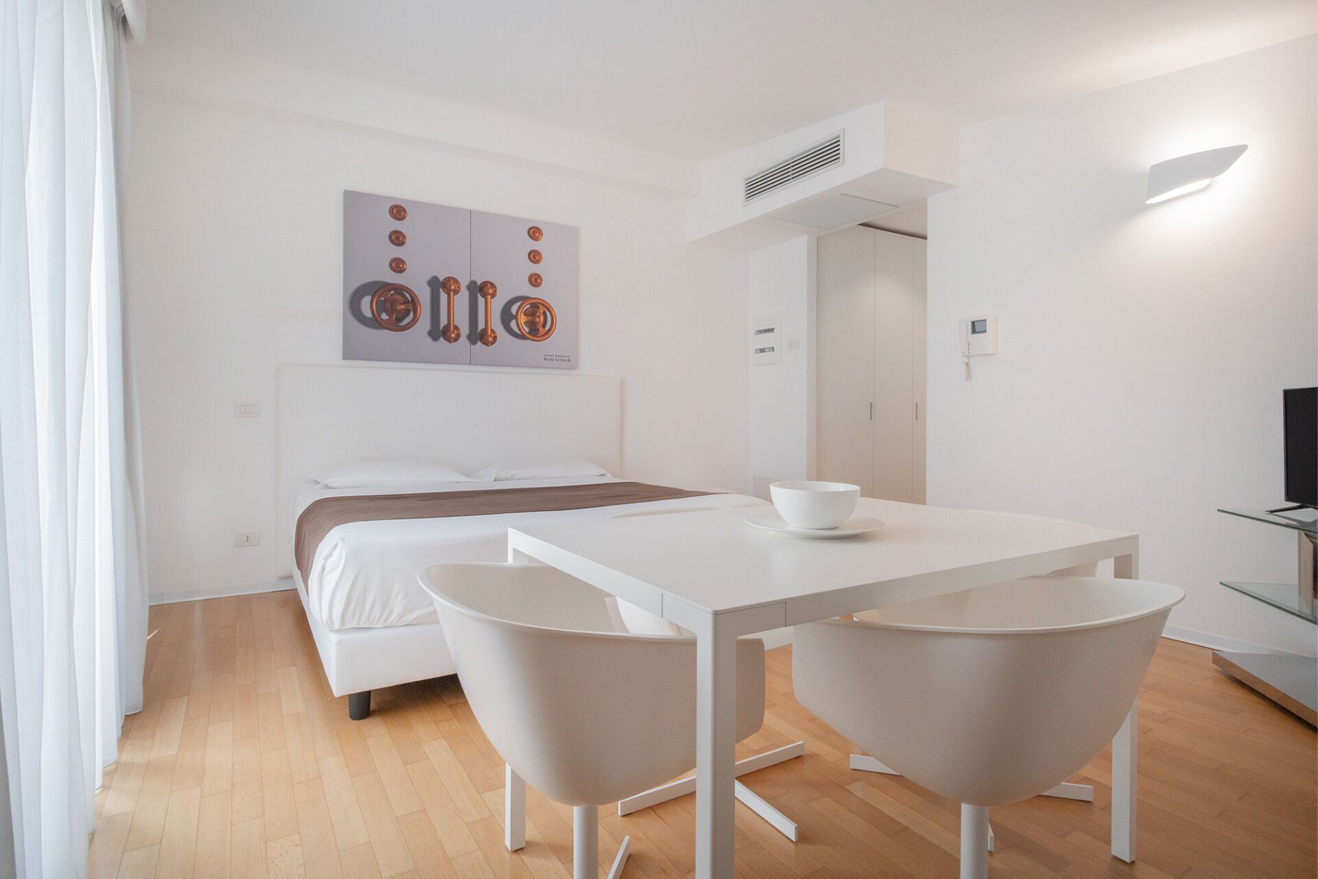 A modern hotel room with a bed, a white table with two chairs, a white bowl, and a painting on the wall.