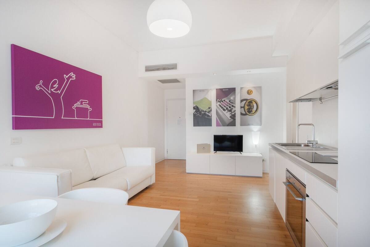 A modern apartment with a white sofa, a dining table with a bowl, a kitchen with a sink and stove, and a living area with a TV and artwork.