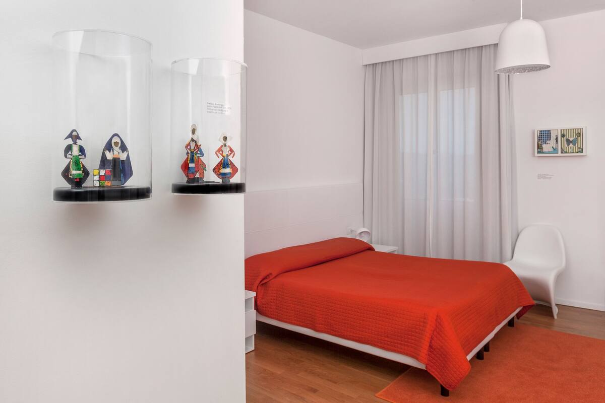 A modern bedroom with a bed, white walls, a window with curtains, and a white chair.