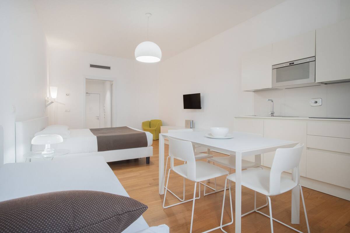 A modern, minimalist hotel room with a bed, a dining area with a table and chairs, a kitchen with a microwave, and a TV mounted on the wall.