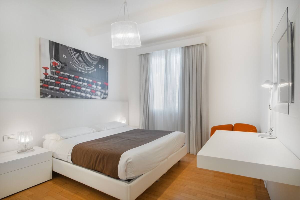 A modern hotel room with a bed, bedside table, and a desk with a chair.